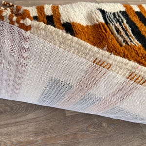 Puede incluir: Una alfombra enrollada con un respaldo de malla blanca. La alfombra presenta un patrón de formas geométricas naranjas, negras y blancas. Se ven borlas trenzadas en el borde. La alfombra está sobre una superficie de madera.