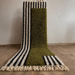Puede incluir: Una alfombra hecha a mano con un diseño único. La alfombra presenta secciones de color verde oliva y crema con rayas negras. La alfombra tiene una textura peluda y un borde con flecos. Se muestra contra una pared neutra y un suelo de madera.
