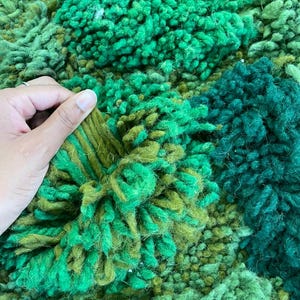 May include: Close-up of a textured rug in shades of green. The rug has a mix of textures, including tufted and looped sections, creating a visually rich and tactile surface. The colour palette ranges from light to dark greens.