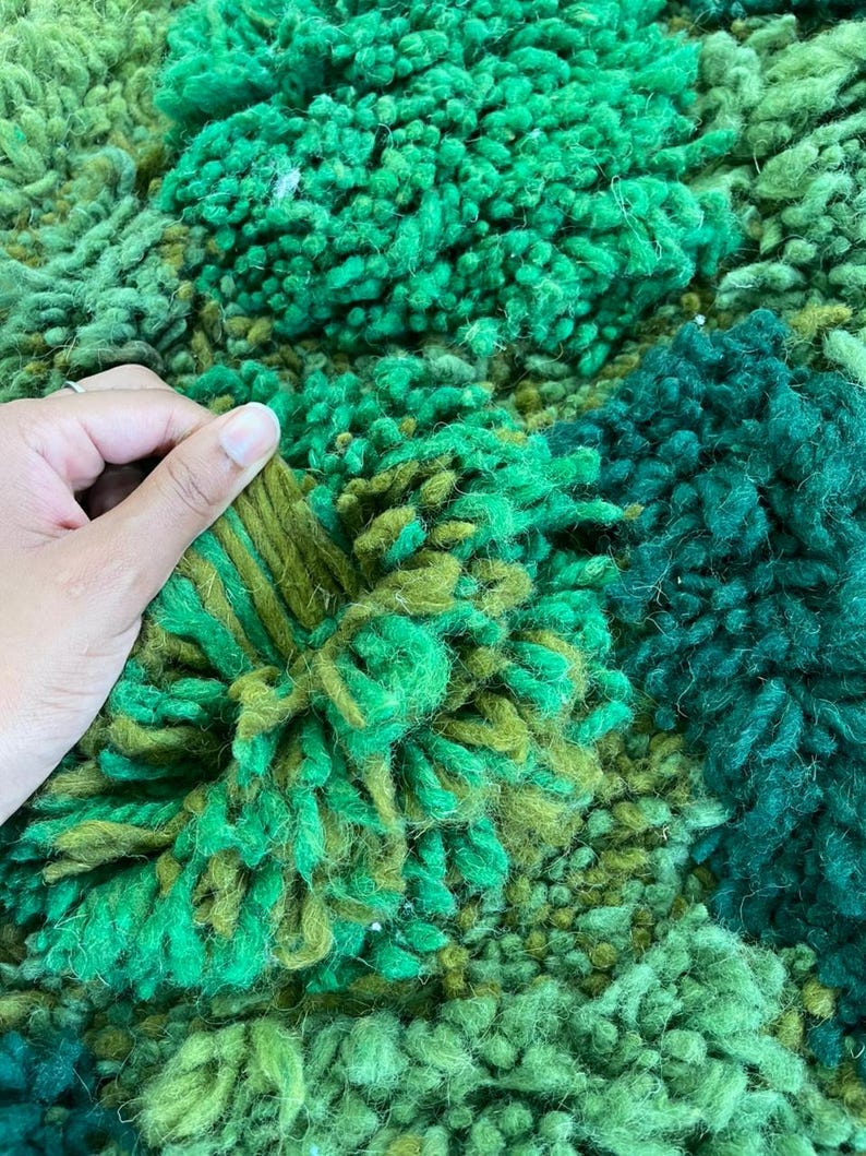 May include: Close-up of a textured rug in shades of green. The rug has a mix of textures, including tufted and looped sections, creating a visually rich and tactile surface. The colour palette ranges from light to dark greens.