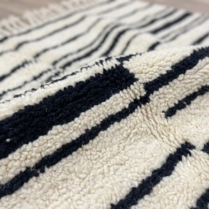May include: A thick, cream-colored wool rug featuring a pattern of bold, black horizontal stripes. The rug has a high-pile, textured surface and includes a fringe detail along the edge, showcasing the soft, knotted construction of the material.