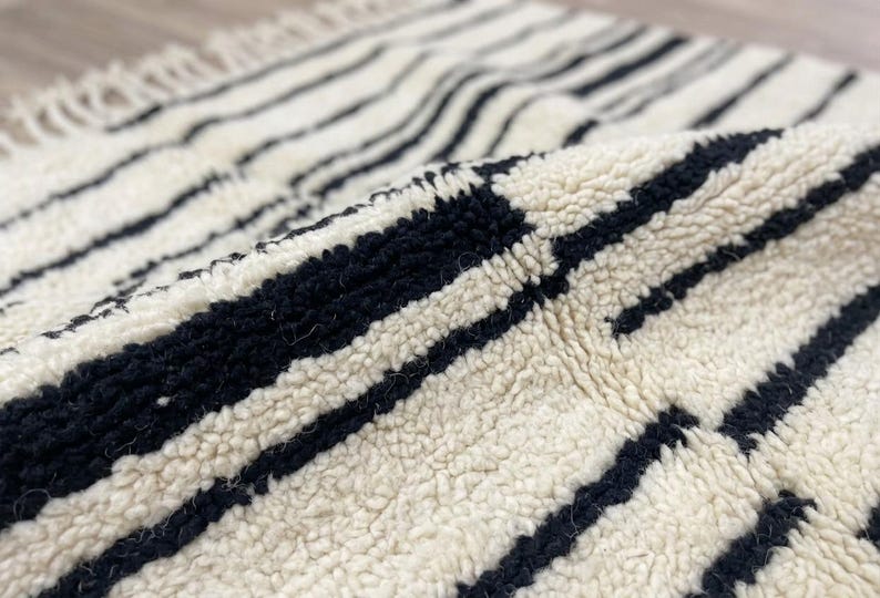 May include: A thick, cream-colored wool rug featuring a pattern of bold, black horizontal stripes. The rug has a high-pile, textured surface and includes a fringe detail along the edge, showcasing the soft, knotted construction of the material.