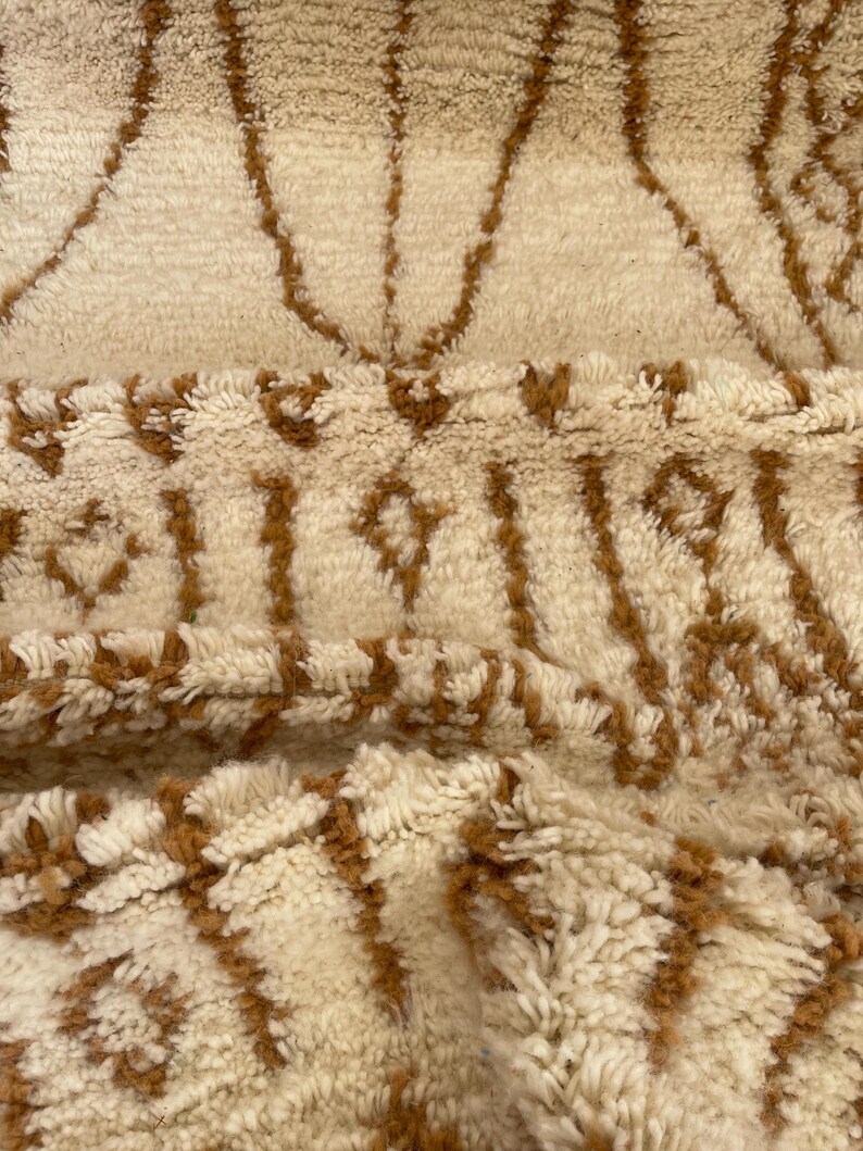 May include: Close-up of a cream-coloured rug with a geometric pattern in shades of brown. The rug has a thick, textured pile, and the design features vertical and diamond-shaped elements. The rug appears to be made of natural fibres.
