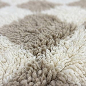 May include: Close-up of a textured rug with a repeating diamond pattern. The rug features alternating diamonds of off-white and light brown, creating a soft, inviting texture. The fibers are thick and plush.