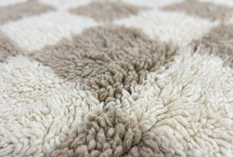 May include: Close-up of a textured rug with a repeating diamond pattern. The rug features alternating diamonds of off-white and light brown, creating a soft, inviting texture. The fibers are thick and plush.