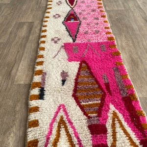 Op de afbeelding: Een lang, rechthoekig tapijt met een witte achtergrond en een geometrisch patroon in roze, rood en bruin. Het tapijt heeft aan beide uiteinden franjes.