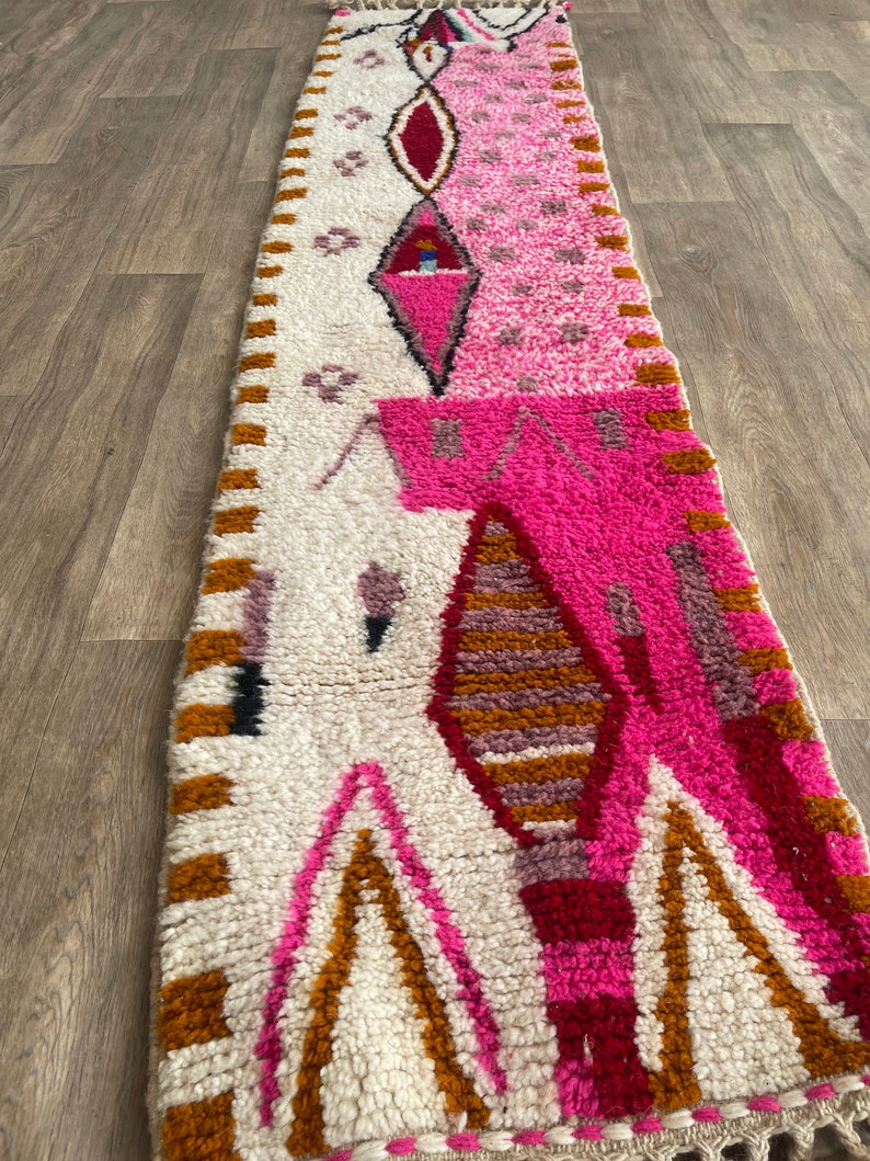 Op de afbeelding: Een lang, rechthoekig tapijt met een witte achtergrond en een geometrisch patroon in roze, rood en bruin. Het tapijt heeft aan beide uiteinden franjes.