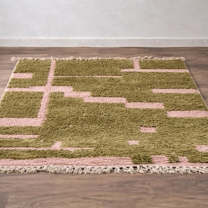 Puede incluir: Una alfombra rectangular con un dise&ntilde;o geom&eacute;trico. La alfombra presenta una combinaci&oacute;n de colores verde oliva y rosa claro. El dise&ntilde;o consta de formas rectangulares y lineales. La alfombra tiene flecos en el borde inferior.