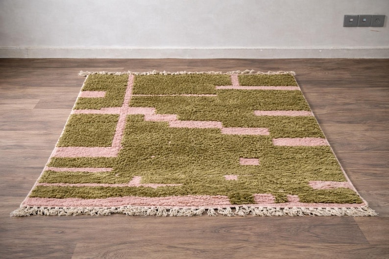 Puede incluir: Una alfombra rectangular con un dise&ntilde;o geom&eacute;trico. La alfombra presenta una combinaci&oacute;n de colores verde oliva y rosa claro. El dise&ntilde;o consta de formas rectangulares y lineales. La alfombra tiene flecos en el borde inferior.