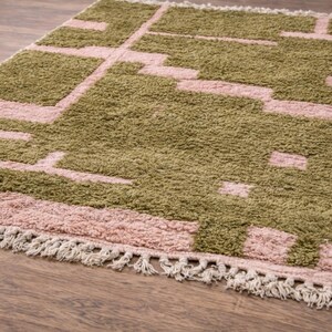 Puede incluir: Una alfombra rectangular con un patr&oacute;n geom&eacute;trico en tonos verde oliva y rosa empolvado. La alfombra tiene una textura afelpada y un borde con flecos. El dise&ntilde;o presenta formas rectangulares y lineales, creando una est&eacute;tica moderna.