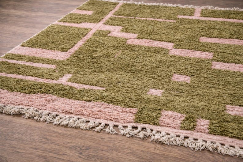 Puede incluir: Una alfombra rectangular con un patr&oacute;n geom&eacute;trico en tonos verde oliva y rosa empolvado. La alfombra tiene una textura afelpada y un borde con flecos. El dise&ntilde;o presenta formas rectangulares y lineales, creando una est&eacute;tica moderna.