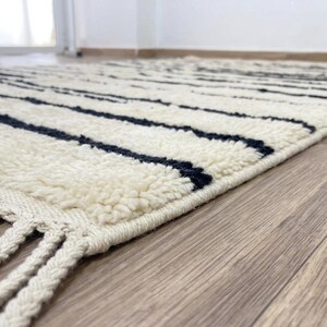 May include: A rectangular area rug featuring a thick, cream-colored high-pile texture with horizontal black stripes. The rug is finished with long, braided cream-colored fringe tassels along the edge, creating a distinct linear pattern.