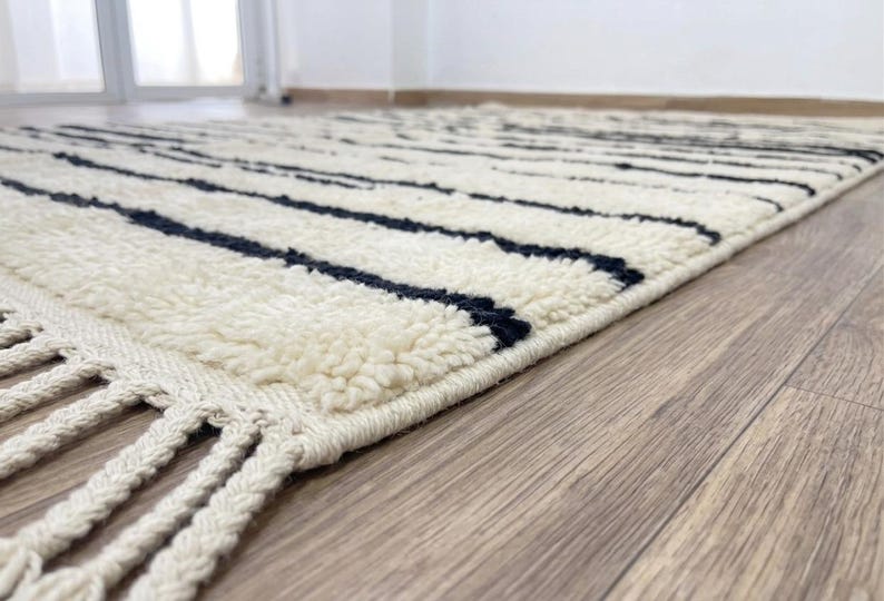 May include: A rectangular area rug featuring a thick, cream-colored high-pile texture with horizontal black stripes. The rug is finished with long, braided cream-colored fringe tassels along the edge, creating a distinct linear pattern.