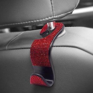 May include: Red rhinestone car seat headrest hook. The hook is made of black plastic and has a red rhinestone design. It is designed to hang on the back of a car seat.
