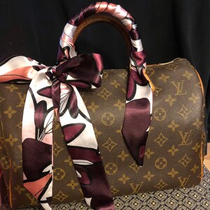May include: A brown leather handbag with a monogram pattern. The handbag has a brown leather handle wrapped with a pink and burgundy floral patterned silk scarf.