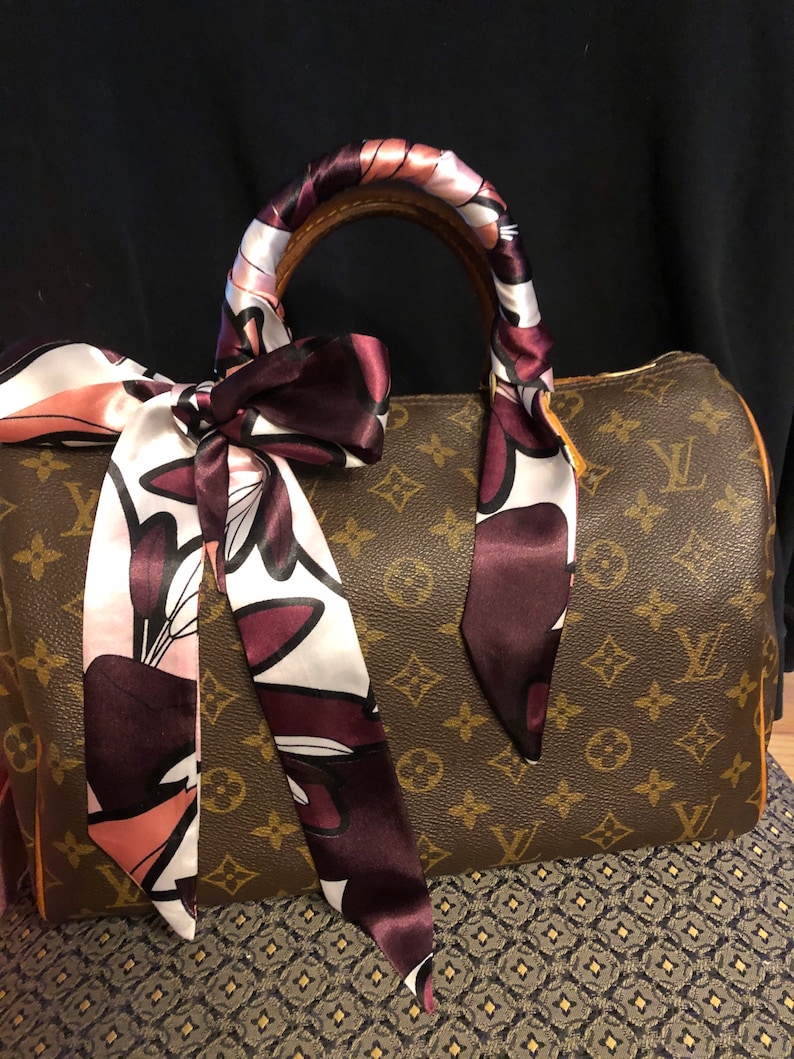 May include: A brown leather handbag with a monogram pattern. The handbag has a brown leather handle wrapped with a pink and burgundy floral patterned silk scarf.