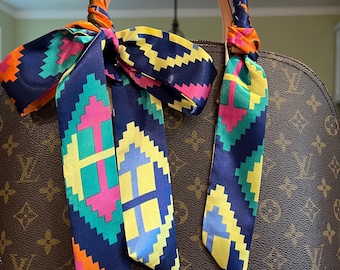 May include: A brown handbag with a light pink base and a patterned design. The handles are wrapped with a colorful scarf featuring a geometric pattern in shades of blue, yellow, pink, and orange. The scarf is tied in a bow.