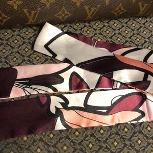 May include: A close-up of a silk scarf with a floral print in shades of pink, white, and burgundy. The scarf is folded and layered, showing the intricate details of the design.
