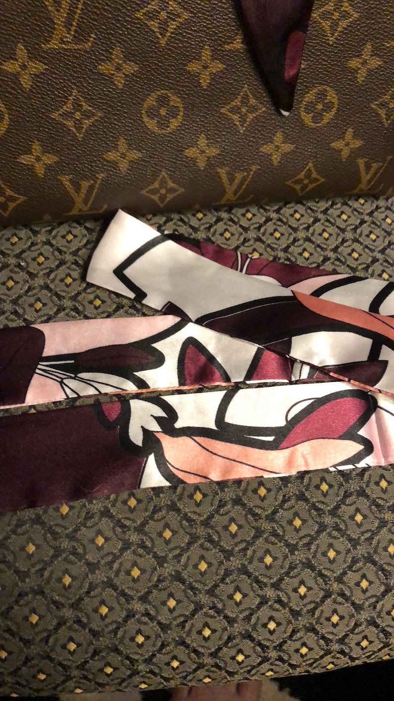 May include: A close-up of a silk scarf with a floral print in shades of pink, white, and burgundy. The scarf is folded and layered, showing the intricate details of the design.