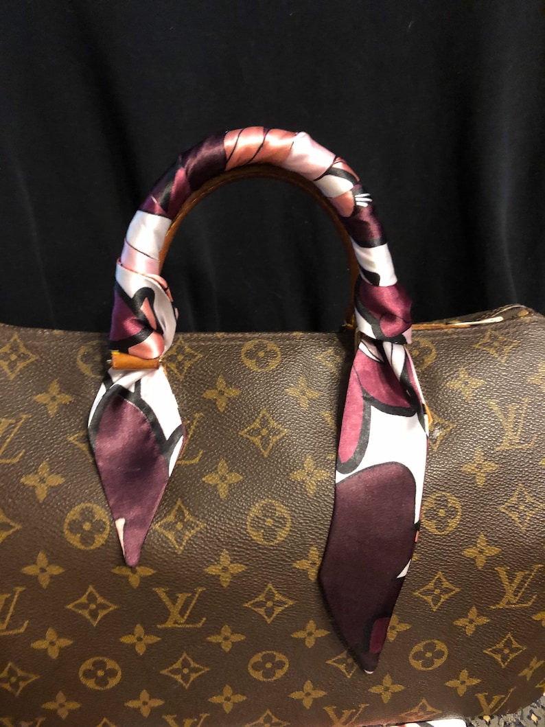 May include: A brown leather handbag with a monogram pattern. The handbag has a brown leather handle with a pink, white, and burgundy patterned scarf tied around it.