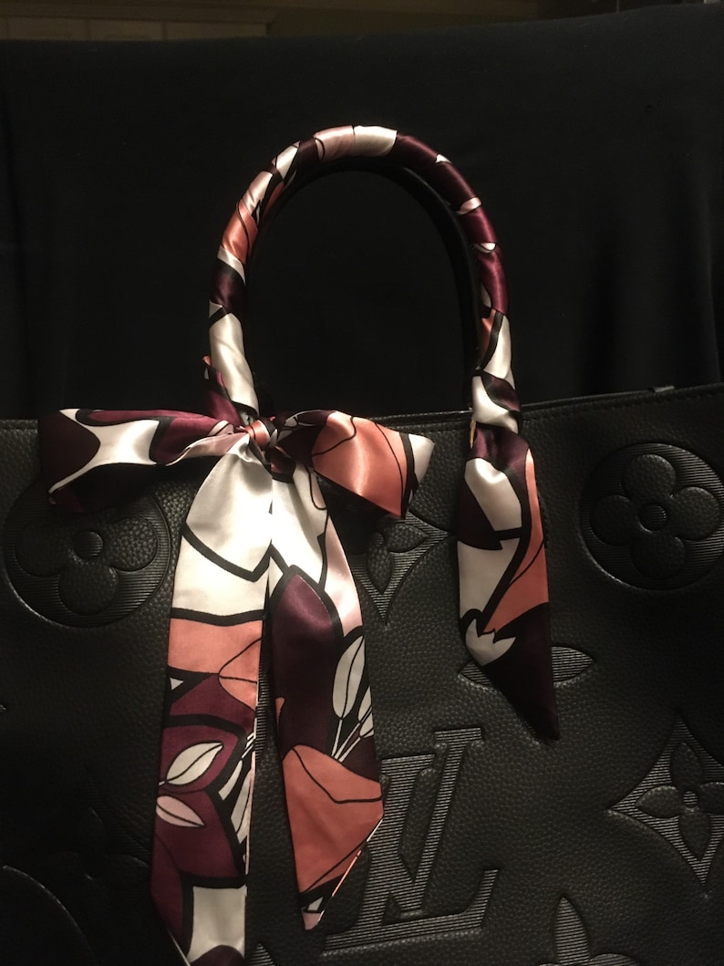 May include: A black leather handbag with a floral patterned scarf tied around the handles. The scarf is in shades of pink, white, and burgundy.