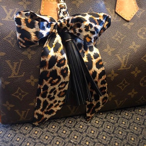 Pode incluir: Um lenço de seda com estampa de leopardo marrom e preto com um tassel de couro preto preso a um chaveiro dourado. O lenço está amarrado em um laço e está pendurado em uma bolsa de couro marrom.