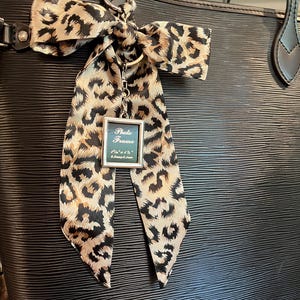 Pode incluir: Uma bolsa tote de couro preto com uma fita de estampa de leopardo amarrada ao redor da alça. Um chaveiro de moldura de foto prateada pendura na fita. A moldura de foto tem as palavras "Photo Frame" escritas nela.