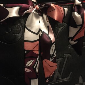 May include: Black leather tote bag with a floral patterned silk scarf tied around the handles. The scarf is in shades of pink, white, and purple.