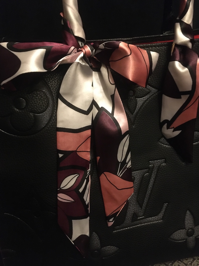May include: Black leather tote bag with a floral patterned silk scarf tied around the handles. The scarf is in shades of pink, white, and purple.