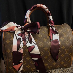May include: A brown and tan monogrammed handbag with a brown leather handle. The handle is wrapped with a pink and burgundy patterned silk scarf.