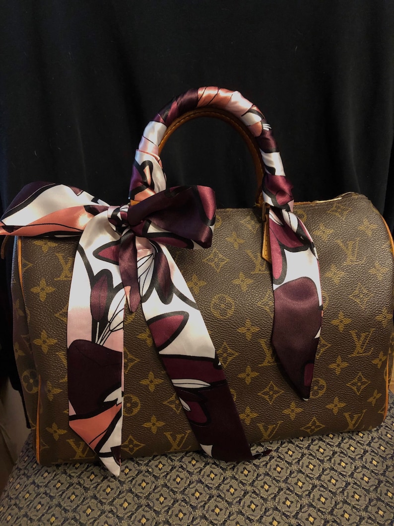 May include: A brown and tan monogrammed handbag with a brown leather handle. The handle is wrapped with a pink and burgundy patterned silk scarf.