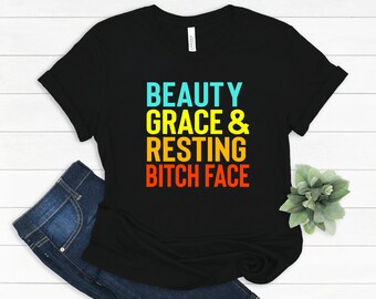 May include: Black t-shirt with the text "BEAUTY GRACE & RESTING BITCH FACE" in turquoise, yellow, and orange. The shirt is paired with blue jeans on a white wooden surface.