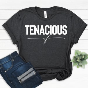 May include: Dark gray t-shirt with the word "TENACIOUS" in bold white letters, with "af" written below in a cursive font. The shirt is paired with blue jeans. The shirt is folded at the sleeves.