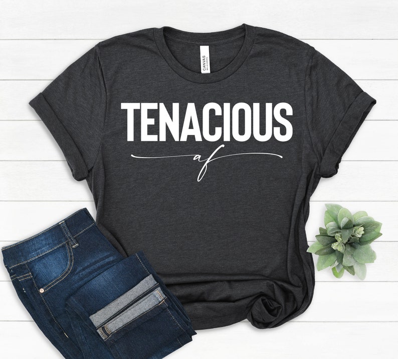 May include: Dark gray t-shirt with the word "TENACIOUS" in bold white letters, with "af" written below in a cursive font. The shirt is paired with blue jeans. The shirt is folded at the sleeves.