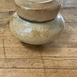 May include: A two-piece ceramic pottery set with a light brown glaze. The top piece is a smaller, rounded lid, and the bottom is a larger, rounded base. The pottery has a rustic, handmade appearance.