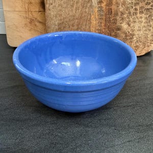 May include: A round, medium-sized ceramic mixing bowl in a solid, vibrant cornflower blue color. The bowl features a smooth, glossy finish with three distinct horizontal ridges embossed around the exterior circumference near the base.