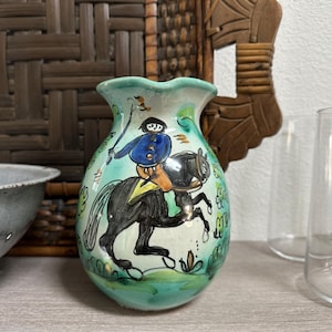 Könnte beinhalten: Handbemalte Keramikvase mit türkisfarbenem Boden und gerüschtem Rand. Die Vase zeigt eine Illustration eines schwarzen Pferdes und Reiters in Blau, Orange und Schwarz. Die Vase ist ein dekorativer Gegenstand.
