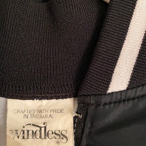 May include: A black windbreaker jacket with white stripes on the sleeves and collar. The jacket has a zip fastening and a label that reads "Crafted with pride in the U.S.A." and "windless".