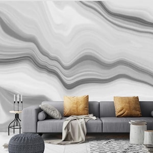 Könnte beinhalten: Ein graues Sofa mit zwei senfgelben Zierkissen und einer hellgrauen Decke. Links steht ein kleiner Beistelltisch aus schwarzem Metall mit drei Kerzen. Ein dunkelgrauer, strukturierter Sitzhocker steht im Vordergrund.