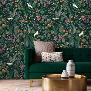 K&ouml;nnte beinhalten: Ein gr&uuml;nes Samtsofa mit zwei Kissen steht vor einer gr&uuml;nen Tapete mit Blumenmuster. Ein runder goldener Couchtisch mit geh&auml;mmerter Oberfl&auml;che steht vor dem Sofa. Zwei wei&szlig;e Keramikvasen stehen auf dem Tisch.