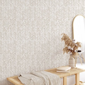 Könnte beinhalten: Eine weiße Wand mit einer beigefarbener Zickzackmuster-Tapete. Im Vordergrund befindet sich eine Holzbank mit einer beigefarbenen Wolldecke. Rechts im Bild steht eine Vase mit getrocknetem Pampasgras.
