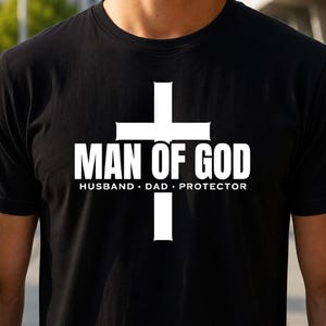 May include: A black short-sleeved t-shirt featuring a large white cross graphic. The text "MAN OF GOD" is printed in bold white capital letters across the center of the cross, with the words "HUSBAND • DAD • PROTECTOR" printed below.