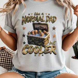 Peut inclure: Un t-shirt gris clair avec un motif de glacière camouflage remplie de glace et de canettes. Le texte "like a NORMAL DAD but COOLER" est écrit en lettres camouflage, entouré d'étoiles marron et vert foncé autour du motif central.
