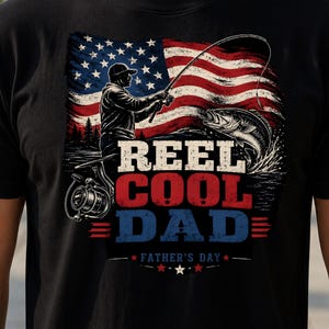 Peut inclure: T-shirt noir avec un motif sur le thème de la pêche. Le design comprend un drapeau américain, un pêcheur, un poisson et le texte "REEL COOL DAD - FÊTE DES PÈRES". Le graphisme est en rouge, blanc et bleu.