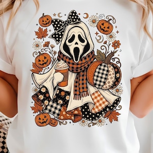 Peut inclure: Un t-shirt blanc avec une illustration graphique d'un personnage fantôme portant un nœud à pois noir et blanc, une écharpe à carreaux et tenant une tasse de café. Le motif inclut des citrouilles orange, des feuilles d'automne et des marguerites.
