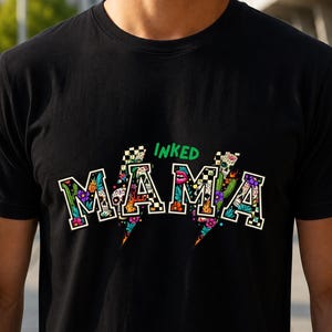 Peut inclure: Un t-shirt noir avec le mot MAMA écrit en grandes lettres de style bloc remplies de motifs colorés incluant des crânes, des fleurs, des cactus et des damiers. Le mot INKED est imprimé en vert au-dessus des lettres, avec des formes d'éclairs.