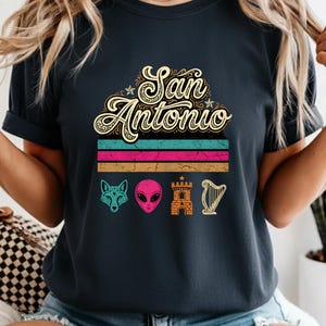 Peut inclure: Un t-shirt gris anthracite avec le texte San Antonio écrit dans une police crème stylisée. Sous le texte se trouvent trois bandes horizontales turquoise, magenta et beige. Quatre icônes figurent en bas : un renard turquoise, une tête d'alien rose, un château beige et une harpe beige.