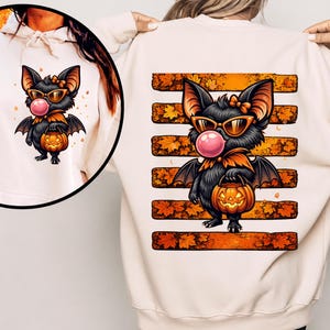 Peut inclure: Un sweat-shirt crème arborant une illustration de chauve-souris noire portant des lunettes de soleil orange et faisant une bulle rose. La chauve-souris tient un seau citrouille devant des bandes horizontales orange ornées de feuilles d'automne.
