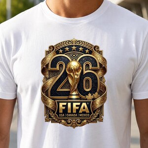 Peut inclure: Un t-shirt blanc à manches courtes arborant un motif central représentant le nombre 26 en or et noir, intégrant un trophée doré, cinq étoiles et le texte FIFA USA | CANADA | MEXICO dans un cadre rectangulaire doré ornementé.