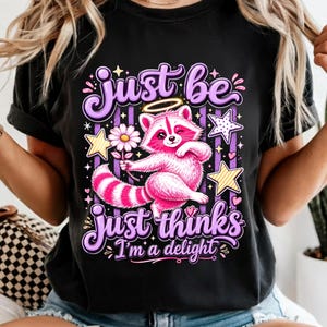 May include: A black t-shirt featuring a graphic of a pink raccoon with a halo holding a daisy. The design includes purple text that reads Just be, Just thinks, and I'm a delight, surrounded by yellow and white stars and purple vertical stripes.